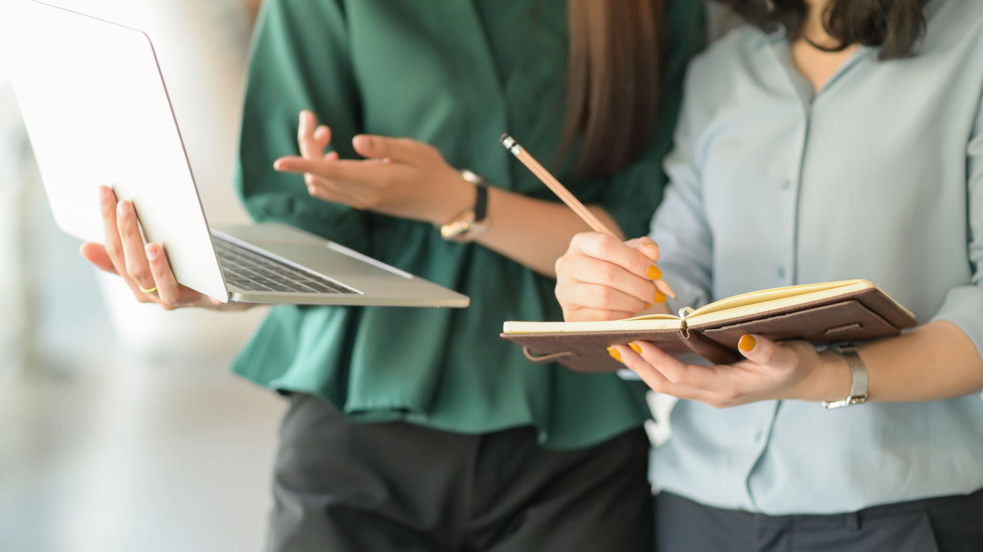 Uma mulher mostrando a tela do notebook para outra mulher que está tomando nota em um caderno