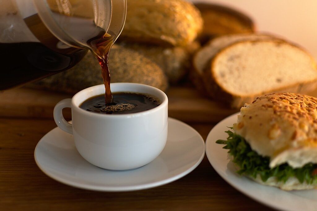 Café sendo colocado na xícara com vários pães ao redor.
