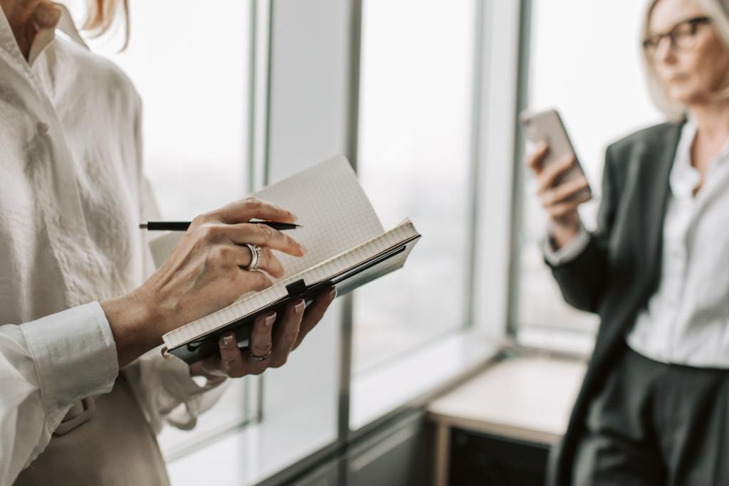 Mulher fazendo anotações em um caderno e outra mulher mexendo no celular.